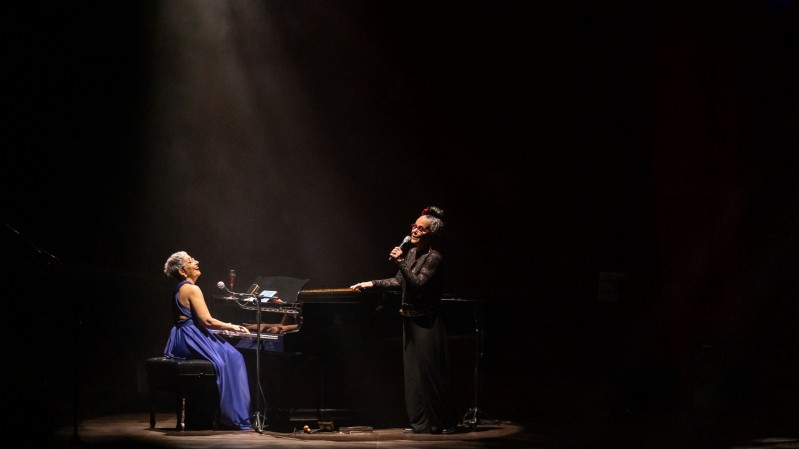 Em um palco escuro iluminado por um feixe de luz vindo do alto, duas artistas se apresentam ao lado de um piano de cauda. Uma delas est&aacute; sentada ao instrumento, usando um vestido azul longo, enquanto a outra permanece de p&eacute;, segurando um microfone. A ilumina&ccedil;&atilde;o concentra a aten&ccedil;&atilde;o na dupla e cria um forte contraste com o fundo escuro do teatro. O piano ocupa o centro da cena, cercado por microfones e equipamentos de som, refor&ccedil;ando o ambiente de apresenta&ccedil;&atilde;o musical ao vivo.