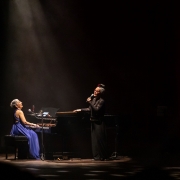 Em um palco escuro iluminado por um feixe de luz vindo do alto, duas artistas se apresentam ao lado de um piano de cauda. Uma delas est&aacute; sentada ao instrumento, usando um vestido azul longo, enquanto a outra permanece de p&eacute;, segurando um microfone. A ilumina&ccedil;&atilde;o concentra a aten&ccedil;&atilde;o na dupla e cria um forte contraste com o fundo escuro do teatro. O piano ocupa o centro da cena, cercado por microfones e equipamentos de som, refor&ccedil;ando o ambiente de apresenta&ccedil;&atilde;o musical ao vivo.