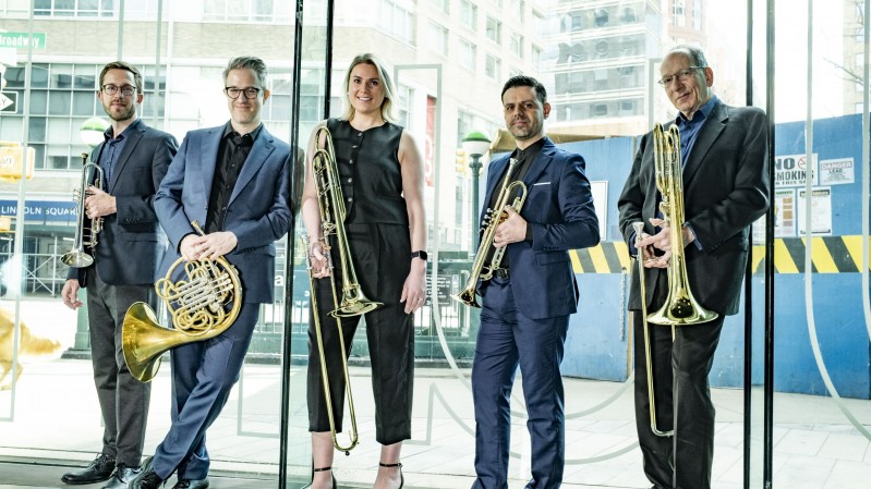 A imagem mostra os cinco m&uacute;sicos do American Brass Quintet posando em p&eacute; em um ambiente interno com grandes paredes de vidro, tendo ao fundo uma paisagem urbana com pr&eacute;dios e estruturas de constru&ccedil;&atilde;o vis&iacute;veis do lado de fora. As cinco pessoas est&atilde;o alinhadas lado a lado, voltadas para a c&acirc;mera. Cada pessoa segura um instrumento de metal da fam&iacute;lia dos metais de sopro, predominantemente trombones e um trompa (French horn). As roupas s&atilde;o formais ou semiformais, em tons escuros: Quatro pessoas usam ternos ou conjuntos sociais em azul-marinho, preto ou cinza. Uma pessoa ao centro veste um conjunto preto sem mangas. Os instrumentos possuem acabamento dourado brilhante, refletindo a luz ambiente. O local parece ser o sagu&atilde;o ou entrada de um edif&iacute;cio moderno, com amplas superf&iacute;cies de vidro.