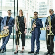 A imagem mostra os cinco m&uacute;sicos do American Brass Quintet posando em p&eacute; em um ambiente interno com grandes paredes de vidro, tendo ao fundo uma paisagem urbana com pr&eacute;dios e estruturas de constru&ccedil;&atilde;o vis&iacute;veis do lado de fora. As cinco pessoas est&atilde;o alinhadas lado a lado, voltadas para a c&acirc;mera. Cada pessoa segura um instrumento de metal da fam&iacute;lia dos metais de sopro, predominantemente trombones e um trompa (French horn). As roupas s&atilde;o formais ou semiformais, em tons escuros: Quatro pessoas usam ternos ou conjuntos sociais em azul-marinho, preto ou cinza. Uma pessoa ao centro veste um conjunto preto sem mangas. Os instrumentos possuem acabamento dourado brilhante, refletindo a luz ambiente. O local parece ser o sagu&atilde;o ou entrada de um edif&iacute;cio moderno, com amplas superf&iacute;cies de vidro.