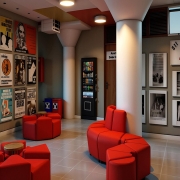 A imagem mostra o interior de um espa&ccedil;o cultural, com paredes decoradas por cartazes e fotografias relacionadas ao teatro. &Agrave; esquerda, h&aacute; uma grande composi&ccedil;&atilde;o de p&ocirc;steres de diferentes espet&aacute;culos, em preto e branco e coloridos. No centro, aparecem poltronas e pufes vermelhos, dispostos em uma &aacute;rea de conviv&ecirc;ncia, al&eacute;m de uma m&aacute;quina de bebidas. &Agrave; direita, a parede re&uacute;ne fotografias emolduradas de apresenta&ccedil;&otilde;es e o letreiro &ldquo;Teatro de Arena&rdquo;. O ambiente tem piso claro, colunas brancas, ilumina&ccedil;&atilde;o quente e decora&ccedil;&atilde;o inspirada na hist&oacute;ria do teatro.