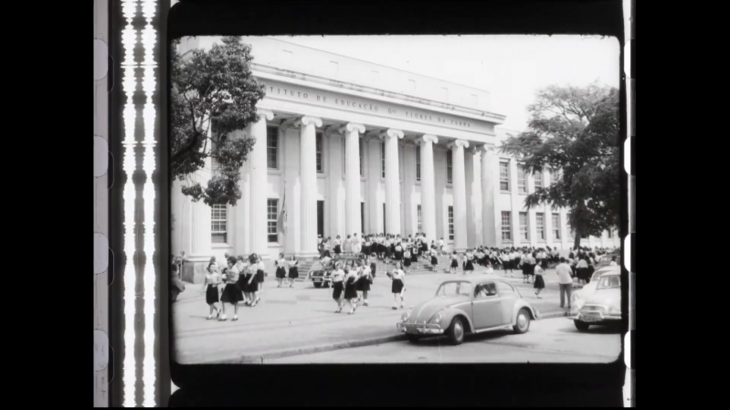 Fotografia em preto e branco de um grande pr&eacute;dio neocl&aacute;ssico com uma extensa fileira de colunas na fachada, onde se l&ecirc; parcialmente a inscri&ccedil;&atilde;o "Instituto de Educa&ccedil;&atilde;o". Em frente ao edif&iacute;cio, uma multid&atilde;o de estudantes uniformizadas, vestindo saias escuras e blusas claras, desce as escadarias e caminha pela rua. Carros antigos, incluindo um fusca, est&atilde;o estacionados ou em movimento na via, refor&ccedil;ando o clima retr&ocirc; da cena.