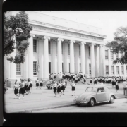 Fotografia em preto e branco de um grande pr&eacute;dio neocl&aacute;ssico com uma extensa fileira de colunas na fachada, onde se l&ecirc; parcialmente a inscri&ccedil;&atilde;o "Instituto de Educa&ccedil;&atilde;o". Em frente ao edif&iacute;cio, uma multid&atilde;o de estudantes uniformizadas, vestindo saias escuras e blusas claras, desce as escadarias e caminha pela rua. Carros antigos, incluindo um fusca, est&atilde;o estacionados ou em movimento na via, refor&ccedil;ando o clima retr&ocirc; da cena.