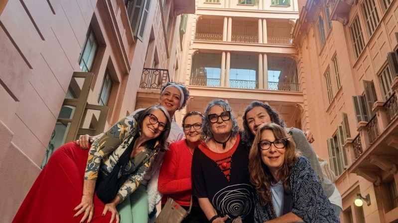 Em uma pra&ccedil;a interna cercada por edif&iacute;cios de v&aacute;rios andares com arquitetura cl&aacute;ssica, seis pessoas posam juntas para a c&acirc;mera em um enquadramento feito de baixo para cima. As pessoas est&atilde;o reunidas no centro da composi&ccedil;&atilde;o, inclinadas em dire&ccedil;&atilde;o &agrave; lente, ocupando diferentes n&iacute;veis do espa&ccedil;o. Os figurinos combinam pe&ccedil;as em tons de vermelho, azul, verde, preto e estampas variadas. Ao fundo, destacam-se fachadas em tons claros, varandas, janelas com venezianas e colunas que se elevam at&eacute; os andares superiores. A ilumina&ccedil;&atilde;o natural valoriza a arquitetura do local e cria uma atmosfera acolhedora, destacando o grupo no centro da cena.