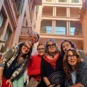 Em uma pra&ccedil;a interna cercada por edif&iacute;cios de v&aacute;rios andares com arquitetura cl&aacute;ssica, seis pessoas posam juntas para a c&acirc;mera em um enquadramento feito de baixo para cima. As pessoas est&atilde;o reunidas no centro da composi&ccedil;&atilde;o, inclinadas em dire&ccedil;&atilde;o &agrave; lente, ocupando diferentes n&iacute;veis do espa&ccedil;o. Os figurinos combinam pe&ccedil;as em tons de vermelho, azul, verde, preto e estampas variadas. Ao fundo, destacam-se fachadas em tons claros, varandas, janelas com venezianas e colunas que se elevam at&eacute; os andares superiores. A ilumina&ccedil;&atilde;o natural valoriza a arquitetura do local e cria uma atmosfera acolhedora, destacando o grupo no centro da cena.