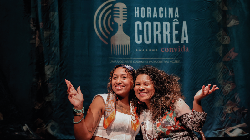 Imagem mostra duas mulheres negras sorrindo e posando juntas diante de um painel azul-escuro. A mulher &agrave; esquerda &eacute; a cantora Cristal. Ela usa blusa branca, acess&oacute;rios nos bra&ccedil;os e um len&ccedil;o estampado no pesco&ccedil;o; a mulher &agrave; direita &eacute; a cantora P&acirc;mela Amaro. Ela veste uma blusa estampada e tem cabelos longos e cacheados. As duas fazem gestos com as m&atilde;os. Ao fundo, h&aacute; a ilustra&ccedil;&atilde;o de um microfone e o texto &ldquo;Horacina Corr&ecirc;a convida&rdquo;, acompanhado da frase &ldquo;Uma voz abre caminho para outras vozes&rdquo;.