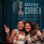 Imagem mostra duas mulheres negras sorrindo e posando juntas diante de um painel azul-escuro. A mulher &agrave; esquerda &eacute; a cantora Cristal. Ela usa blusa branca, acess&oacute;rios nos bra&ccedil;os e um len&ccedil;o estampado no pesco&ccedil;o; a mulher &agrave; direita &eacute; a cantora P&acirc;mela Amaro. Ela veste uma blusa estampada e tem cabelos longos e cacheados. As duas fazem gestos com as m&atilde;os. Ao fundo, h&aacute; a ilustra&ccedil;&atilde;o de um microfone e o texto &ldquo;Horacina Corr&ecirc;a convida&rdquo;, acompanhado da frase &ldquo;Uma voz abre caminho para outras vozes&rdquo;.