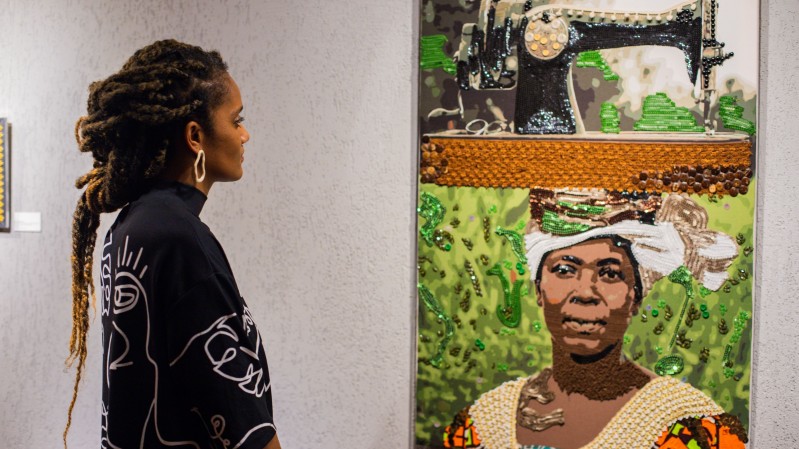 A imagem mostra o interior de uma galeria ou espa&ccedil;o de exposi&ccedil;&atilde;o de arte. &Agrave; esquerda, h&aacute; uma pessoa de perfil observando uma obra exposta na parede. A pessoa veste uma pe&ccedil;a de roupa escura de mangas compridas com desenhos lineares claros estampados e usa um brinco geom&eacute;trico. Os cabelos est&atilde;o presos em longas mechas. &Agrave; direita, encontra-se uma obra de arte vertical composta por diferentes elementos e texturas. Na parte superior da obra h&aacute; a representa&ccedil;&atilde;o de uma m&aacute;quina de costura antiga. Abaixo dela, aparece o retrato de uma pessoa usando um tecido ou turbante branco na cabe&ccedil;a e uma vestimenta clara. O fundo do retrato &eacute; predominantemente verde, com detalhes que parecem ter sido feitos com materiais brilhantes ou tridimensionais, criando relevo e textura. A moldura ou divis&atilde;o entre as partes da obra apresenta uma faixa horizontal em tom alaranjado, tamb&eacute;m com apar&ecirc;ncia texturizada. O conjunto parece ser uma t&eacute;cnica mista, combinando pintura com materiais aplicados sobre a superf&iacute;cie.