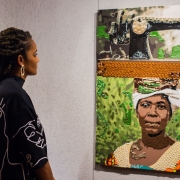 A imagem mostra o interior de uma galeria ou espa&ccedil;o de exposi&ccedil;&atilde;o de arte. &Agrave; esquerda, h&aacute; uma pessoa de perfil observando uma obra exposta na parede. A pessoa veste uma pe&ccedil;a de roupa escura de mangas compridas com desenhos lineares claros estampados e usa um brinco geom&eacute;trico. Os cabelos est&atilde;o presos em longas mechas. &Agrave; direita, encontra-se uma obra de arte vertical composta por diferentes elementos e texturas. Na parte superior da obra h&aacute; a representa&ccedil;&atilde;o de uma m&aacute;quina de costura antiga. Abaixo dela, aparece o retrato de uma pessoa usando um tecido ou turbante branco na cabe&ccedil;a e uma vestimenta clara. O fundo do retrato &eacute; predominantemente verde, com detalhes que parecem ter sido feitos com materiais brilhantes ou tridimensionais, criando relevo e textura. A moldura ou divis&atilde;o entre as partes da obra apresenta uma faixa horizontal em tom alaranjado, tamb&eacute;m com apar&ecirc;ncia texturizada. O conjunto parece ser uma t&eacute;cnica mista, combinando pintura com materiais aplicados sobre a superf&iacute;cie.