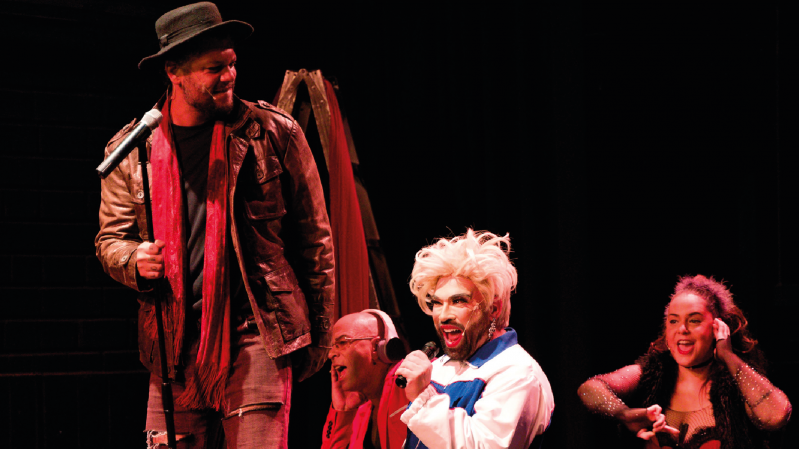 A imagem mostra quatro atores em cena, durante uma apresenta&ccedil;&atilde;o teatral ou musical, diante de um fundo preto. &Agrave; esquerda, um homem usa chap&eacute;u, jaqueta de couro marrom, camisa escura e uma longa echarpe vermelha, segurando um microfone. Ao centro, uma pessoa com peruca branca volumosa e maquiagem marcante canta ao microfone, vestindo uma roupa branca com detalhes azuis e vermelhos. Ao fundo, outra pessoa aparece sentada, parcialmente encoberta, tamb&eacute;m segurando um microfone. &Agrave; direita, uma mulher de cabelos escuros e cacheados, com mechas avermelhadas, sorri e se movimenta durante a apresenta&ccedil;&atilde;o. A ilumina&ccedil;&atilde;o destaca os artistas e cria um clima de espet&aacute;culo no palco.