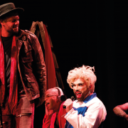 A imagem mostra quatro atores em cena, durante uma apresenta&ccedil;&atilde;o teatral ou musical, diante de um fundo preto. &Agrave; esquerda, um homem usa chap&eacute;u, jaqueta de couro marrom, camisa escura e uma longa echarpe vermelha, segurando um microfone. Ao centro, uma pessoa com peruca branca volumosa e maquiagem marcante canta ao microfone, vestindo uma roupa branca com detalhes azuis e vermelhos. Ao fundo, outra pessoa aparece sentada, parcialmente encoberta, tamb&eacute;m segurando um microfone. &Agrave; direita, uma mulher de cabelos escuros e cacheados, com mechas avermelhadas, sorri e se movimenta durante a apresenta&ccedil;&atilde;o. A ilumina&ccedil;&atilde;o destaca os artistas e cria um clima de espet&aacute;culo no palco.