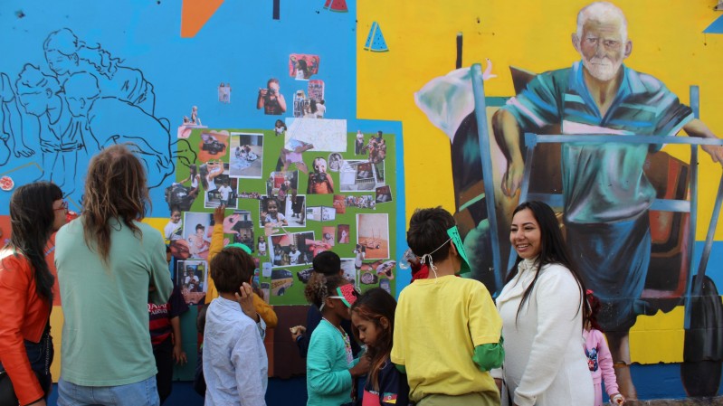 Fotografia de um grupo de crian&ccedil;as e adultos reunidos diante de um mural colorido em um espa&ccedil;o externo. A parede combina &aacute;reas em azul e amarelo e apresenta diferentes elementos visuais, incluindo desenhos, fotografias coladas e a pintura de uma pessoa idosa puxando um ve&iacute;culo de tra&ccedil;&atilde;o humana. No centro da cena, h&aacute; um painel verde preenchido com v&aacute;rias fotografias e imagens que parecem fazer parte de uma atividade coletiva. As crian&ccedil;as observam o painel e interagem entre si, enquanto alguns adultos acompanham a a&ccedil;&atilde;o. 

