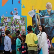 Fotografia de um grupo de crian&ccedil;as e adultos reunidos diante de um mural colorido em um espa&ccedil;o externo. A parede combina &aacute;reas em azul e amarelo e apresenta diferentes elementos visuais, incluindo desenhos, fotografias coladas e a pintura de uma pessoa idosa puxando um ve&iacute;culo de tra&ccedil;&atilde;o humana. No centro da cena, h&aacute; um painel verde preenchido com v&aacute;rias fotografias e imagens que parecem fazer parte de uma atividade coletiva. As crian&ccedil;as observam o painel e interagem entre si, enquanto alguns adultos acompanham a a&ccedil;&atilde;o. 

