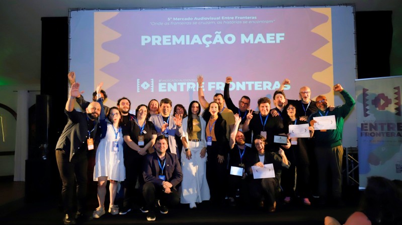 Um grupo numeroso de participantes re&uacute;ne-se sobre um palco para uma fotografia coletiva durante a cerim&ocirc;nia de premia&ccedil;&atilde;o do Mercado Audiovisual Entre Fronteras. Algumas pessoas exibem certificados e credenciais penduradas no pesco&ccedil;o, enquanto outras erguem os bra&ccedil;os. Ao fundo, uma grande tela projetada traz os dizeres &ldquo;Premia&ccedil;&atilde;o MAEF&rdquo; e a marca do &ldquo;5&ordm; Mercado Audiovisual Entre Fronteras&rdquo;. A imagem registra o encerramento de um momento de celebra&ccedil;&atilde;o com representantes de diferentes projetos e iniciativas ligadas ao evento.