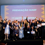 Um grupo numeroso de participantes re&uacute;ne-se sobre um palco para uma fotografia coletiva durante a cerim&ocirc;nia de premia&ccedil;&atilde;o do Mercado Audiovisual Entre Fronteras. Algumas pessoas exibem certificados e credenciais penduradas no pesco&ccedil;o, enquanto outras erguem os bra&ccedil;os. Ao fundo, uma grande tela projetada traz os dizeres &ldquo;Premia&ccedil;&atilde;o MAEF&rdquo; e a marca do &ldquo;5&ordm; Mercado Audiovisual Entre Fronteras&rdquo;. A imagem registra o encerramento de um momento de celebra&ccedil;&atilde;o com representantes de diferentes projetos e iniciativas ligadas ao evento.
