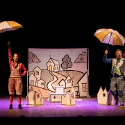 Em um palco iluminado, duas pessoas caracterizadas como personagens de um espet&aacute;culo est&atilde;o em cena segurando guarda-chuvas coloridos. &Agrave; esquerda, uma mulher usa roupa em tons de rosa e vermelho, enquanto, &agrave; direita, um homem veste roupas coloridas e estampadas. Ao centro, h&aacute; um grande painel com o desenho de uma pequena vila, com casas, igreja, &aacute;rvores e um caminho. O cen&aacute;rio tamb&eacute;m conta com pequenas casas de papel espalhadas pelo palco, araras com figurinos e outros objetos c&ecirc;nicos. Ao fundo, predomina um fundo preto.