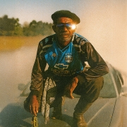 Pessoa agachada sobre o capô de um automóvel em uma estrada de terra coberta por poeira iluminada pelo sol baixo. A pessoa usa boina escura, óculos escuros refletivos, camisa do Grêmio nas cores azul, preto e branco, calças escuras e botas. Uma das mãos segura um pingente dourado. A luz do sol incide diretamente sobre os óculos, produzindo um reflexo brilhante. Ao fundo, a paisagem é composta por árvores desfocadas e uma atmosfera dourada criada pela poeira suspensa no ar.