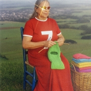 Pessoa de cabelos grisalhos sentada em uma cadeira azul em meio a uma paisagem de campo verde. A pessoa veste um longo vestido vermelho com o número 7 estampado em branco na parte frontal e usa óculos escuros que refletem a luz do sol, criando um brilho intenso sobre uma das lentes. Nas mãos, segura agulhas de tricô e trabalha em uma peça de lã verde fluorescente. Ao lado, há um cesto de vime com várias peças de tricô dobradas em cores vibrantes, como rosa, azul, amarelo e lilás. Ao fundo, aparecem colinas e áreas de vegetação desfocadas sob luz natural suave.



