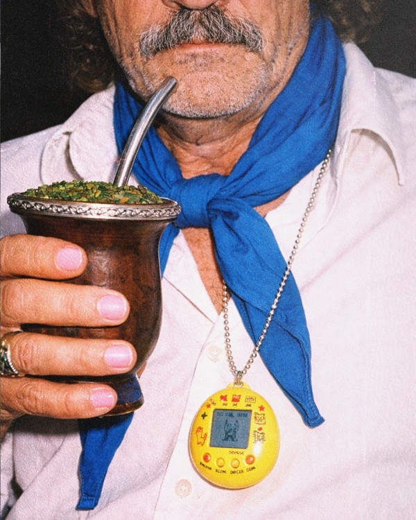 Close no tronco de uma pessoa vestindo camisa branca e lenço azul amarrado ao pescoço. Em uma das mãos, a pessoa segura uma cuia de chimarrão de madeira com erva-mate e bomba metálica. Sobre o peito, presa a uma corrente prateada, há um dispositivo eletrônico amarelo semelhante a um brinquedo virtual portátil, com uma pequena tela e botões na parte inferior. A fotografia destaca os detalhes dos objetos e das roupas, com fundo escuro e iluminação direta concentrada nos elementos centrais da cena.