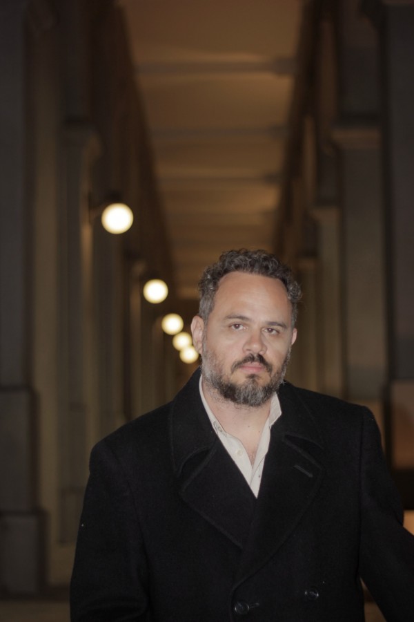 Foto de André Hernandez, um homem branco, de barba e cabelo grisalhos. Ele aparece do peito para cima, veste blazer preto e camisa bege e olha para a esquerda da câmera. É noite, e André está em um local externo, à frente de diversas pilastras