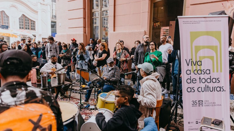 A foto mostra uma roda de samba. H&aacute; muitas pessoas na foto, dan&ccedil;ando e tocando instrumentos. Em primeiro plano, aparece um cartaz de divulga&ccedil;&atilde;o da evento com a frase "Viva a casa de todas as culturas".