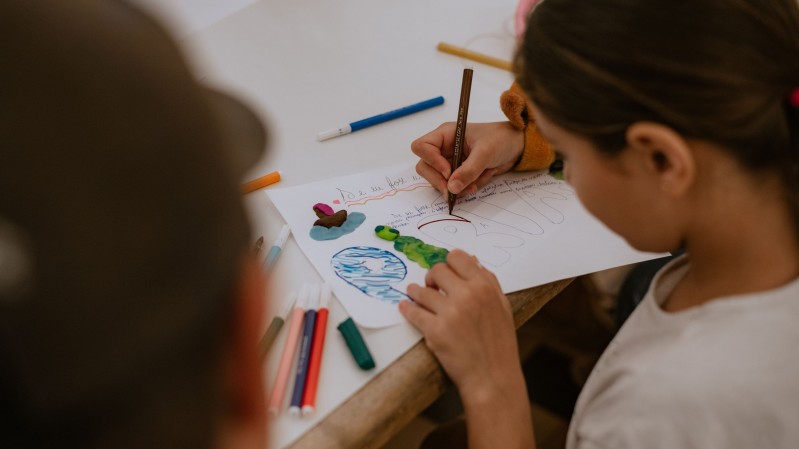Uma crian&ccedil;a est&aacute; sentada &agrave; mesa enquanto desenha e escreve em uma folha de papel utilizando uma caneta azul. Sobre a folha h&aacute; desenhos coloridos e elementos feitos com massa de modelar em tons de verde, azul, marrom e rosa. Diversos canet&otilde;es e peda&ccedil;os de massinha est&atilde;o distribu&iacute;dos sobre a mesa, incluindo algumas embalagens pl&aacute;sticas abertas. A fotografia foi feita de cima, destacando a atividade criativa e os materiais utilizados.