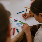 Uma crian&ccedil;a est&aacute; sentada &agrave; mesa enquanto desenha e escreve em uma folha de papel utilizando uma caneta azul. Sobre a folha h&aacute; desenhos coloridos e elementos feitos com massa de modelar em tons de verde, azul, marrom e rosa. Diversos canet&otilde;es e peda&ccedil;os de massinha est&atilde;o distribu&iacute;dos sobre a mesa, incluindo algumas embalagens pl&aacute;sticas abertas. A fotografia foi feita de cima, destacando a atividade criativa e os materiais utilizados.