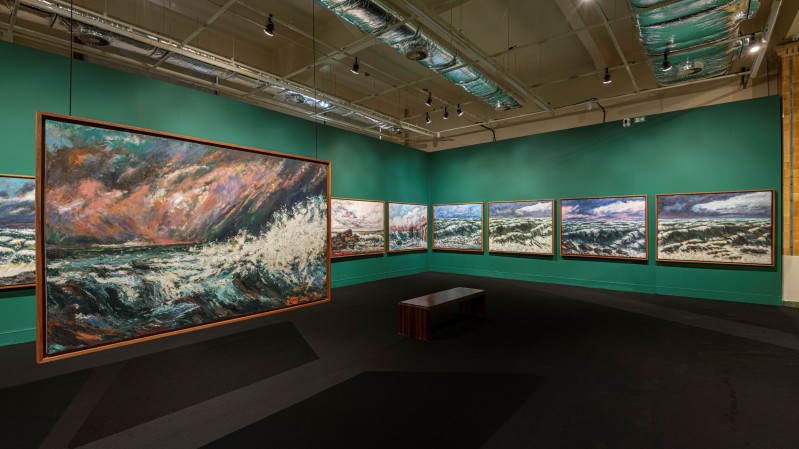 A imagem mostra o interior de uma galeria de arte do MARGS. O ambiente &eacute; amplo, com paredes pintadas em um tom de verde-turquesa escuro e piso escuro, criando um contraste marcante com as obras expostas. arede esquerda, destaca-se uma grande pintura de dimens&otilde;es monumentais, posicionada perpendicularmente &agrave; parede, em primeiro plano. A obra principal apresenta uma cena mar&iacute;tima dram&aacute;tica, com ondas agitadas, espuma branca e um c&eacute;u composto por pinceladas expressivas em tons de azul, cinza, rosa e alaranjado. Ao longo das paredes do fundo e da direita h&aacute; uma sequ&ecirc;ncia de quadros retangulares emoldurados, aparentemente pertencentes &agrave; mesma s&eacute;rie tem&aacute;tica. As pinturas retratam paisagens mar&iacute;timas ou costeiras, com predomin&acirc;ncia de azuis, brancos e cinzas. No centro da sala h&aacute; um banco retangular de madeira escura, destinado &agrave; contempla&ccedil;&atilde;o das obras. O teto &eacute; alto e revela parte da infraestrutura do edif&iacute;cio, incluindo dutos de ventila&ccedil;&atilde;o met&aacute;licos aparentes e trilhos de ilumina&ccedil;&atilde;o. N&atilde;o h&aacute; visitantes vis&iacute;veis no ambiente, o que transmite uma sensa&ccedil;&atilde;o de tranquilidade e foco nas obras.