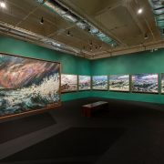 A imagem mostra o interior de uma galeria de arte do MARGS. O ambiente &eacute; amplo, com paredes pintadas em um tom de verde-turquesa escuro e piso escuro, criando um contraste marcante com as obras expostas. arede esquerda, destaca-se uma grande pintura de dimens&otilde;es monumentais, posicionada perpendicularmente &agrave; parede, em primeiro plano. A obra principal apresenta uma cena mar&iacute;tima dram&aacute;tica, com ondas agitadas, espuma branca e um c&eacute;u composto por pinceladas expressivas em tons de azul, cinza, rosa e alaranjado. Ao longo das paredes do fundo e da direita h&aacute; uma sequ&ecirc;ncia de quadros retangulares emoldurados, aparentemente pertencentes &agrave; mesma s&eacute;rie tem&aacute;tica. As pinturas retratam paisagens mar&iacute;timas ou costeiras, com predomin&acirc;ncia de azuis, brancos e cinzas. No centro da sala h&aacute; um banco retangular de madeira escura, destinado &agrave; contempla&ccedil;&atilde;o das obras. O teto &eacute; alto e revela parte da infraestrutura do edif&iacute;cio, incluindo dutos de ventila&ccedil;&atilde;o met&aacute;licos aparentes e trilhos de ilumina&ccedil;&atilde;o. N&atilde;o h&aacute; visitantes vis&iacute;veis no ambiente, o que transmite uma sensa&ccedil;&atilde;o de tranquilidade e foco nas obras.