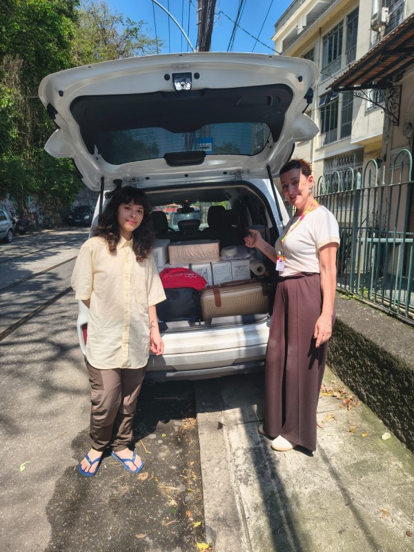 Em uma rua arborizada durante o dia, duas pessoas posam ao lado de um veículo utilitário branco com o porta-malas aberto. O compartimento está preenchido por caixas, embalagens, malas e outros volumes organizados para transporte. Ao fundo, aparecem edificações residenciais, grades, calçadas e fiação urbana sob céu azul. A fotografia registra um momento relacionado à movimentação e ao deslocamento de materiais, com destaque para o veículo carregado e para os itens acomodados em seu interior.
