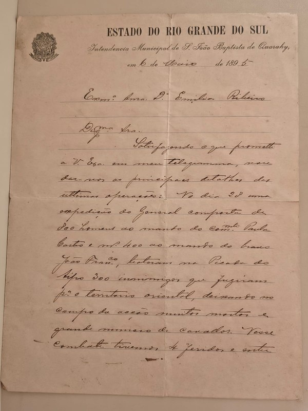 Fotografia de um documento histórico em papel amarelado, apresentado na posição vertical sobre uma superfície clara. O documento possui o cabeçalho impresso “Estado do Rio Grande do Sul” e “Intendência Municipal de S. João Baptista de Quarahy”, acompanhado do brasão da República no canto superior esquerdo. A data registrada é 6 de maio de 1895. A maior parte da página é ocupada por texto manuscrito em tinta escura, escrito em caligrafia cursiva e distribuído em linhas horizontais. O papel apresenta marcas de dobra ao centro, pequenas manchas e sinais de envelhecimento compatíveis com sua antiguidade. A imagem destaca tanto os elementos impressos quanto a escrita manual, evidenciando o caráter histórico e documental da correspondência.
