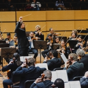 Imagem mostra a orquestra sinf&ocirc;nica durante uma apresenta&ccedil;&atilde;o em um audit&oacute;rio. Dezenas de m&uacute;sicos, vestidos de preto, est&atilde;o sentados em cadeiras e tocam instrumentos de cordas, como violinos, violas e violoncelos, diante de estantes com partituras. Ao centro, o maestro, tamb&eacute;m vestido de preto, est&aacute; de p&eacute; e ergue o bra&ccedil;o para conduzir a orquestra. Ao fundo, as paredes s&atilde;o revestidas com pain&eacute;is de madeira em tons de amarelo e marrom.