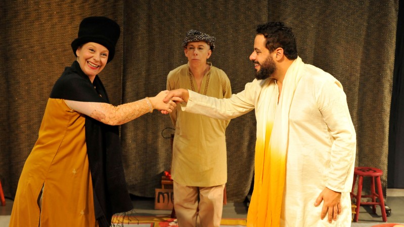 A imagem mostra tr&ecirc;s atores em uma cena que aparenta ser uma apresenta&ccedil;&atilde;o teatral, diante de uma grande cortina escura. Em primeiro plano, &agrave; esquerda, uma pessoa veste roupa longa em tons de amarelo e preto, al&eacute;m de uma cobertura preta arredondada na cabe&ccedil;a. A pessoa estende o bra&ccedil;o direito em dire&ccedil;&atilde;o ao centro da imagem. &Agrave; direita, outra pessoa usa traje branco comprido, com uma faixa ou tecido amarelo na parte frontal, e estende a m&atilde;o esquerda. As m&atilde;os das duas pessoas se encontram no centro da cena. Entre as duas, um pouco mais ao fundo, est&aacute; uma terceira pessoa, vestindo roupa larga em tons de bege e amarelo-claro, cal&ccedil;a clara e uma pe&ccedil;a estampada sobre a cabe&ccedil;a. A pessoa est&aacute; posicionada de frente, com os bra&ccedil;os pr&oacute;ximos ao corpo. Ao fundo h&aacute; uma cortina marrom-escura, que ocupa praticamente toda a extens&atilde;o da imagem. No lado direito, pr&oacute;ximo &agrave; cortina, aparece um banquinho vermelho. No ch&atilde;o, atr&aacute;s das pessoas, tamb&eacute;m s&atilde;o parcialmente vis&iacute;veis alguns objetos utilizados na cena.