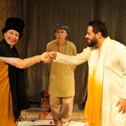 A imagem mostra tr&ecirc;s atores em uma cena que aparenta ser uma apresenta&ccedil;&atilde;o teatral, diante de uma grande cortina escura. Em primeiro plano, &agrave; esquerda, uma pessoa veste roupa longa em tons de amarelo e preto, al&eacute;m de uma cobertura preta arredondada na cabe&ccedil;a. A pessoa estende o bra&ccedil;o direito em dire&ccedil;&atilde;o ao centro da imagem. &Agrave; direita, outra pessoa usa traje branco comprido, com uma faixa ou tecido amarelo na parte frontal, e estende a m&atilde;o esquerda. As m&atilde;os das duas pessoas se encontram no centro da cena. Entre as duas, um pouco mais ao fundo, est&aacute; uma terceira pessoa, vestindo roupa larga em tons de bege e amarelo-claro, cal&ccedil;a clara e uma pe&ccedil;a estampada sobre a cabe&ccedil;a. A pessoa est&aacute; posicionada de frente, com os bra&ccedil;os pr&oacute;ximos ao corpo. Ao fundo h&aacute; uma cortina marrom-escura, que ocupa praticamente toda a extens&atilde;o da imagem. No lado direito, pr&oacute;ximo &agrave; cortina, aparece um banquinho vermelho. No ch&atilde;o, atr&aacute;s das pessoas, tamb&eacute;m s&atilde;o parcialmente vis&iacute;veis alguns objetos utilizados na cena.