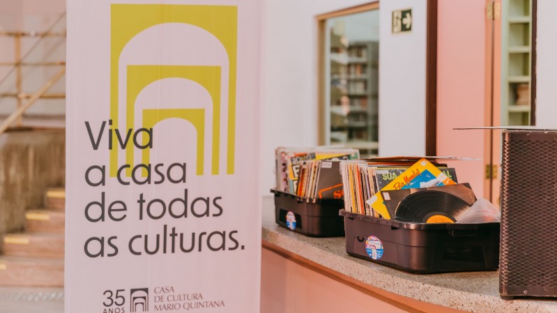 Em um ambiente interno iluminado por luz suave, um banner vertical ocupa o lado esquerdo da imagem com a frase &ldquo;Viva a casa de todas as culturas.&rdquo; e a identifica&ccedil;&atilde;o dos 35 anos da Casa de Cultura Mario Quintana. Ao lado do banner, sobre um balc&atilde;o de superf&iacute;cie clara, est&atilde;o organizadas caixas pl&aacute;sticas contendo dezenas de discos de vinil posicionados na vertical, com capas coloridas vis&iacute;veis. Em primeiro plano, um disco parcialmente exposto destaca-se entre os demais. &Agrave; direita, aparece parte de uma caixa de som de grande porte. Ao fundo, &eacute; poss&iacute;vel observar corredores, uma escadaria e uma porta aberta, compondo um ambiente cultural voltado &agrave; exposi&ccedil;&atilde;o e circula&ccedil;&atilde;o de acervos musicais.