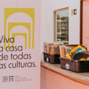 Em um ambiente interno iluminado por luz suave, um banner vertical ocupa o lado esquerdo da imagem com a frase &ldquo;Viva a casa de todas as culturas.&rdquo; e a identifica&ccedil;&atilde;o dos 35 anos da Casa de Cultura Mario Quintana. Ao lado do banner, sobre um balc&atilde;o de superf&iacute;cie clara, est&atilde;o organizadas caixas pl&aacute;sticas contendo dezenas de discos de vinil posicionados na vertical, com capas coloridas vis&iacute;veis. Em primeiro plano, um disco parcialmente exposto destaca-se entre os demais. &Agrave; direita, aparece parte de uma caixa de som de grande porte. Ao fundo, &eacute; poss&iacute;vel observar corredores, uma escadaria e uma porta aberta, compondo um ambiente cultural voltado &agrave; exposi&ccedil;&atilde;o e circula&ccedil;&atilde;o de acervos musicais.