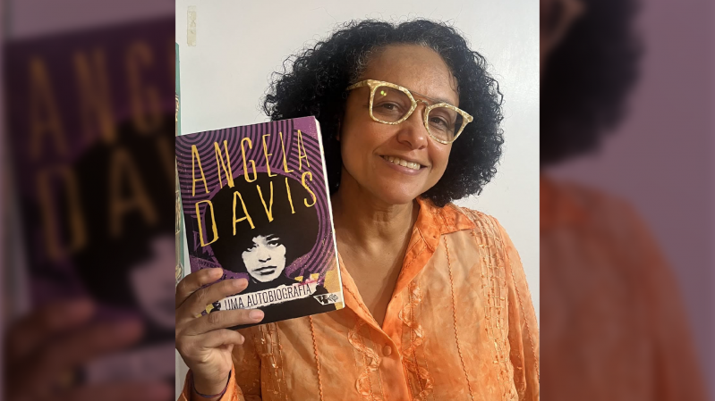 A foto mostra Taiasmin Ohnmacht, uma mulher negra, com cabelos cacheados na altura dos ombros, usando &oacute;culos amarelos e uma camisa laranja. Ela sorri para foto segurando o livro que ser&aacute; debatido no encontro, "Uma Autobiografia" de Angela Davis