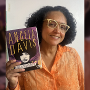 A foto mostra Taiasmin Ohnmacht, uma mulher negra, com cabelos cacheados na altura dos ombros, usando &oacute;culos amarelos e uma camisa laranja. Ela sorri para foto segurando o livro que ser&aacute; debatido no encontro, "Uma Autobiografia" de Angela Davis