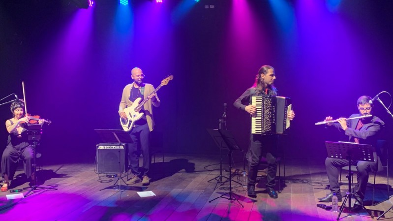 Fotografia de grupo musical se apresentando em um palco amplo e escuro, iluminado por refletores azuis, rosas e violetas posicionados no alto. Quatro m&uacute;sicos est&atilde;o distribu&iacute;dos horizontalmente pelo palco: &agrave; esquerda, uma pessoa toca violino; ao lado, outra toca contrabaixo el&eacute;trico; no centro, uma pessoa executa um acorde&atilde;o; e, &agrave; direita, outra toca flauta transversal. Os m&uacute;sicos est&atilde;o pr&oacute;ximos a estantes de partitura, microfones e amplificadores. O piso de madeira reflete parte da ilumina&ccedil;&atilde;o colorida, criando manchas de luz sobre a superf&iacute;cie. O fundo preto destaca os artistas e os instrumentos, conferindo protagonismo &agrave; apresenta&ccedil;&atilde;o musical e &agrave; composi&ccedil;&atilde;o do conjunto no palco.