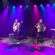 Fotografia de grupo musical se apresentando em um palco amplo e escuro, iluminado por refletores azuis, rosas e violetas posicionados no alto. Quatro m&uacute;sicos est&atilde;o distribu&iacute;dos horizontalmente pelo palco: &agrave; esquerda, uma pessoa toca violino; ao lado, outra toca contrabaixo el&eacute;trico; no centro, uma pessoa executa um acorde&atilde;o; e, &agrave; direita, outra toca flauta transversal. Os m&uacute;sicos est&atilde;o pr&oacute;ximos a estantes de partitura, microfones e amplificadores. O piso de madeira reflete parte da ilumina&ccedil;&atilde;o colorida, criando manchas de luz sobre a superf&iacute;cie. O fundo preto destaca os artistas e os instrumentos, conferindo protagonismo &agrave; apresenta&ccedil;&atilde;o musical e &agrave; composi&ccedil;&atilde;o do conjunto no palco.