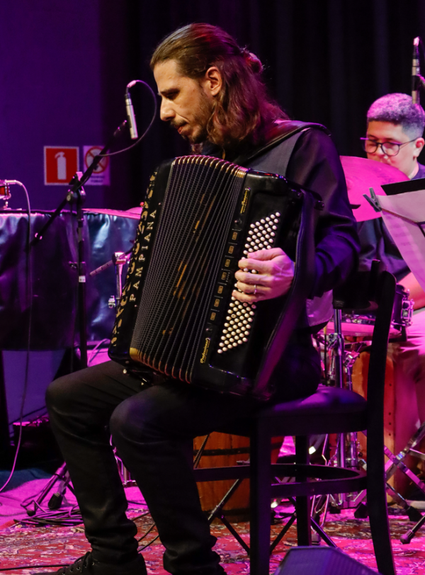 Fotografia de apresentação musical em ambiente fechado com iluminação cênica em tons de roxo e rosa. Em primeiro plano, uma pessoa sentada em uma cadeira toca um acordeão preto de grande porte. O instrumento ocupa o centro da composição, com destaque para os botões brancos e para o fole parcialmente aberto. A pessoa veste roupas escuras e mantém o olhar voltado para o instrumento. Ao fundo, aparecem parte de uma bateria e outra pessoa posicionada atrás dos tambores. Microfones, cabos, suportes e equipamentos de palco completam a cena. O palco possui piso coberto por tapete estampado e cortinas escuras ao fundo, reforçando o ambiente de espetáculo ao vivo.