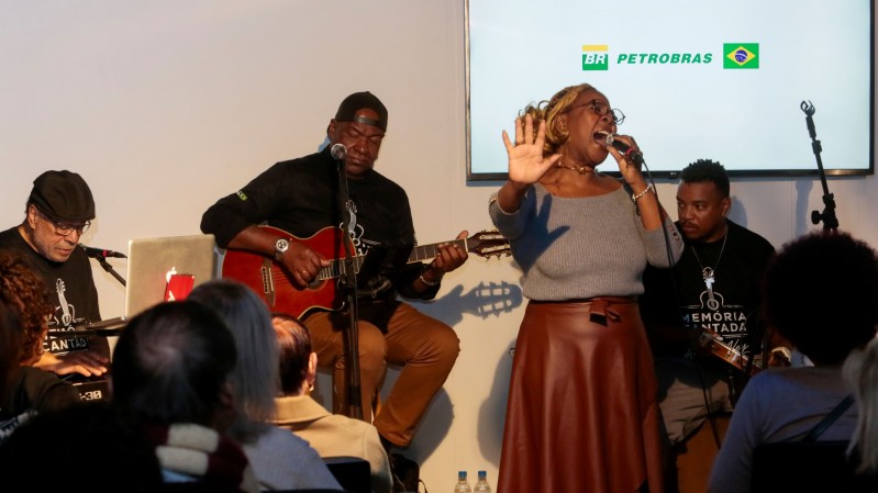 A imagem mostra uma apresenta&ccedil;&atilde;o musical realizada em ambiente interno, diante de um p&uacute;blico sentado. No centro do palco, uma pessoa est&aacute; em p&eacute;, cantando ao microfone e com uma das m&atilde;os levantada. A vestimenta inclui blusa de manga longa em tom cinza e saia marrom. Ao lado, duas pessoas acompanham a apresenta&ccedil;&atilde;o sentadas. Uma delas toca viol&atilde;o ac&uacute;stico de cor avermelhada e est&aacute; posicionada em frente a um microfone. A outra est&aacute; junto a um teclado eletr&ocirc;nico, tamb&eacute;m voltada para o p&uacute;blico. H&aacute; ainda uma quarta pessoa sentada &agrave; direita, pr&oacute;xima aos m&uacute;sicos.