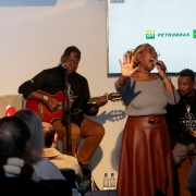 A imagem mostra uma apresenta&ccedil;&atilde;o musical realizada em ambiente interno, diante de um p&uacute;blico sentado. No centro do palco, uma pessoa est&aacute; em p&eacute;, cantando ao microfone e com uma das m&atilde;os levantada. A vestimenta inclui blusa de manga longa em tom cinza e saia marrom. Ao lado, duas pessoas acompanham a apresenta&ccedil;&atilde;o sentadas. Uma delas toca viol&atilde;o ac&uacute;stico de cor avermelhada e est&aacute; posicionada em frente a um microfone. A outra est&aacute; junto a um teclado eletr&ocirc;nico, tamb&eacute;m voltada para o p&uacute;blico. H&aacute; ainda uma quarta pessoa sentada &agrave; direita, pr&oacute;xima aos m&uacute;sicos.